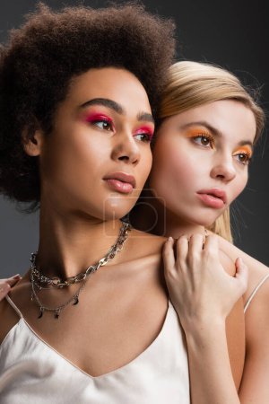Photo for Portrait of pretty multiethnic women with colorful and glossy eye shadows looking away isolated on grey - Royalty Free Image