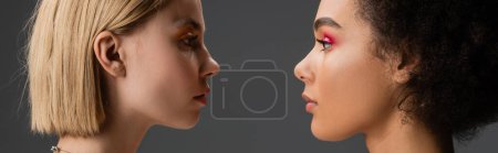 Photo for Side view of interracial blonde and brunette women with makeup looking at each other isolated on grey, banner - Royalty Free Image
