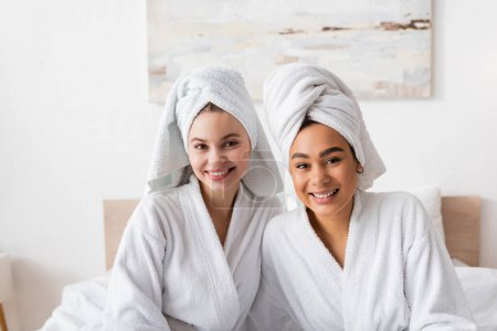 young interracial women in soft bathrobes and towels smiling at camera in bedroom