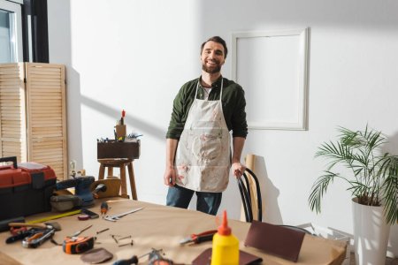 Smiling craftsman in apron standing near blurred tools on table 
