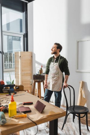 Foto de Positive carpenter standing near chair and tools in workshop - Imagen libre de derechos