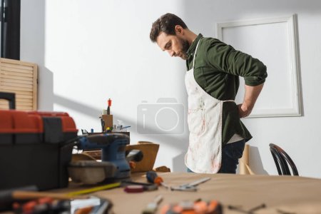 Foto de Side view of bearded carpenter wearing apron near blurred tools in workshop - Imagen libre de derechos