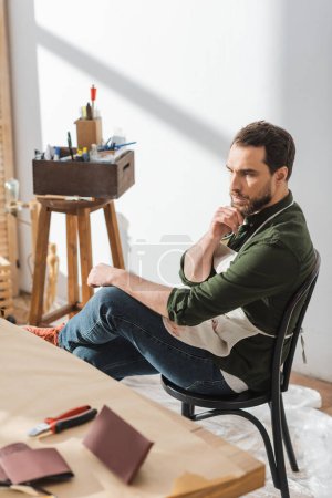 Foto de Pensive craftsman in apron sitting on chair near blurred table in workshop - Imagen libre de derechos