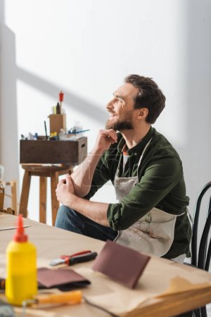 Foto de Smiling carpenter in dirty apron sitting near working table in workshop - Imagen libre de derechos