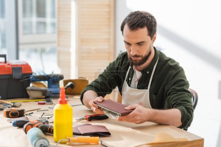 Foto de Bearded carpenter in apron holding sandpaper near tools on table in workshop - Imagen libre de derechos