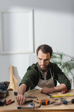 Foto de Craftsman in apron taking tool while working at table in workshop - Imagen libre de derechos