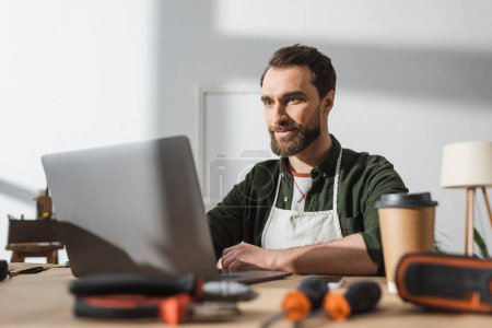 Téléchargez les photos : Bearded carpenter using laptop near blurred takeaway coffee and tools in workshop - en image libre de droit