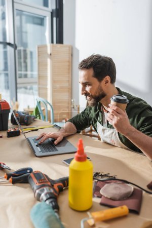 Positive bearded carpenter holding coffee to go and using laptop near tools in workshop 