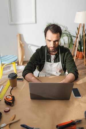 Bärtiger Handwerker in Schürze mit Laptop in der Nähe von Werkzeugen und Kaffee auf dem Tisch in der Werkstatt 