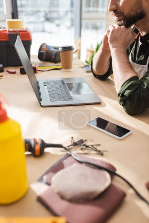 Ausgeschnittene Ansicht eines bärtigen Zimmermanns in Schürze, der in der Nähe von Geräten mit leerem Bildschirm und Geräten auf dem Tisch sitzt 