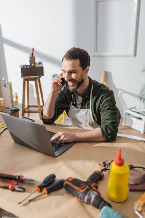 Alegre carpintero hablando en el teléfono inteligente y utilizando el ordenador portátil cerca de herramientas en el taller 