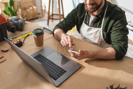 Foto de Cropped view of bearded craftsman using smartphone near laptop and coffee to go in workshop - Imagen libre de derechos
