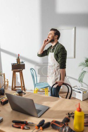 Positive craftsman talking on cellphone near tools and laptop on working table 
