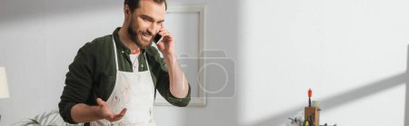 Photo for Smiling carpenter in apron talking on smartphone in workshop, banner - Royalty Free Image