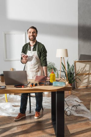 Foto de Smiling carpenter holding smartphone near tools and devices in workshop - Imagen libre de derechos