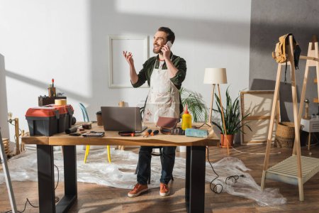Fröhlicher Tischler spricht auf Smartphone neben Werkzeug auf Arbeitstisch im Atelier 
