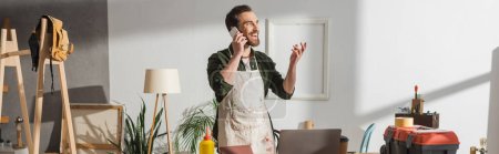 Photo for Smiling craftsman talking on cellphone near tools and laptop in studio, banner - Royalty Free Image