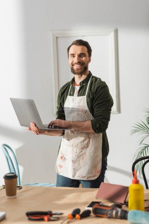Foto de Carpintero positivo en delantal usando portátil cerca de herramientas borrosas y café en taller - Imagen libre de derechos