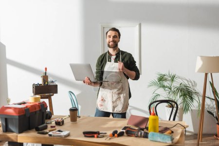 Foto de Smiling carpenter in apron holding laptop and looking at camera near tools in workshop - Imagen libre de derechos