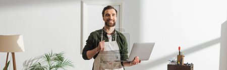 Téléchargez les photos : Cheerful carpenter in apron holding laptop and looking at camera in workshop, banner - en image libre de droit
