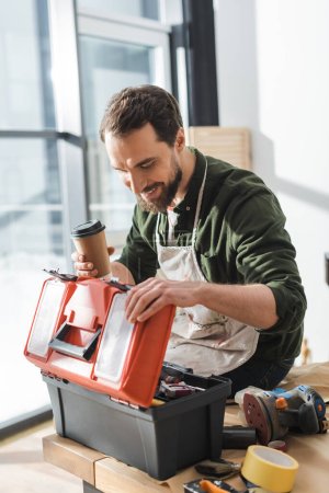 Glücklicher Tischler in Schürze hält Kaffee zum Mitnehmen und öffnet Werkzeugkiste in Werkstatt 
