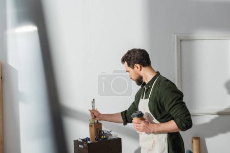 Téléchargez les photos : Vue latérale du menuisier tenant du café à emporter et prenant des pinceaux en atelier - en image libre de droit