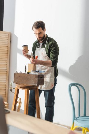 Artesano en delantal sosteniendo pinceles y café para ir en taller 