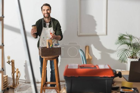 Smiling craftsman holding coffee to go and paintbrushes in workshop 