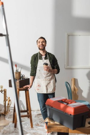 Positive carpenter holding coffee to go and looking at camera in workshop 