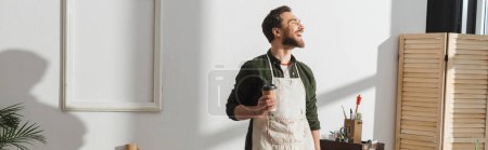 Photo for Smiling bearded repairman in apron holding paper cup in workshop, banner - Royalty Free Image