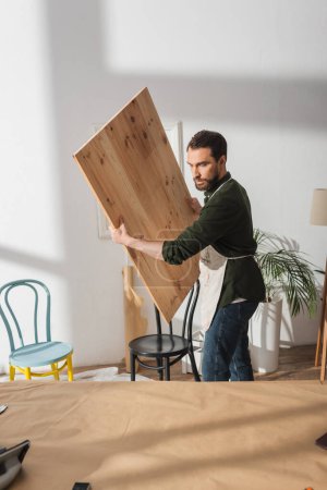 Carpenter holding wooden board near chairs in workshop 
