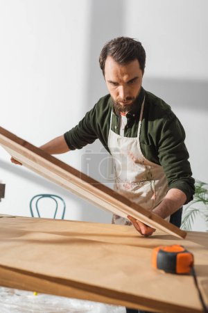 Foto de Artesano barbudo en delantal sosteniendo tablero de madera cerca de regla en taller - Imagen libre de derechos