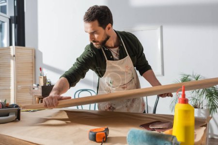 Carpenter in dirty apron putting wooden board on table near ruler and sandpaper 