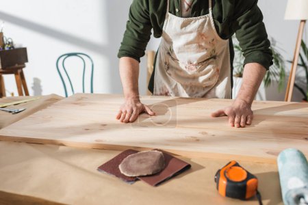 Foto de Vista recortada del trabajador en delantal poniendo tablero de madera en la mesa cerca de regla y papel de lija - Imagen libre de derechos