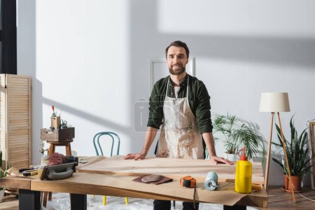 Smiling bearded carpenter in apron looking at camera near wooden board in workshop 