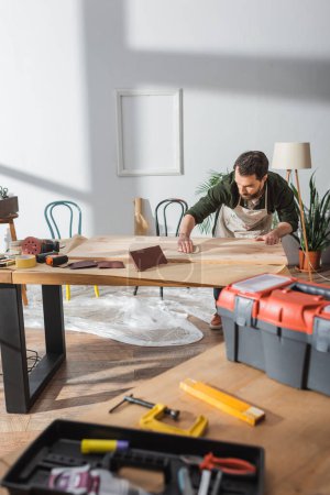 Craftsman sanding wooden board near blurred tools in workshop 