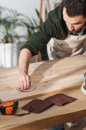 Bearded craftsman sanding wooden board near ruler in workshop 