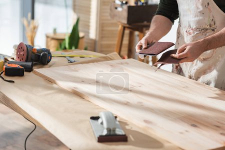 Foto de Cropped view of craftsman holding sandpaper near wooden board and tools - Imagen libre de derechos