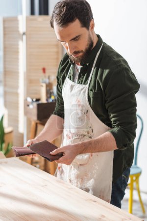 Bearded craftsman in apron holding sandpaper near wooden board 
