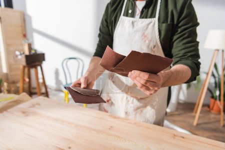 Cropped view of blurred craftsman in apron holding sandpaper while working with wooden board 