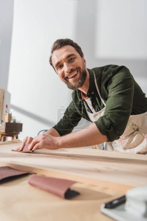 Cheerful bearded carpenter in apron looking at camera while sanding wooden board in workshop 