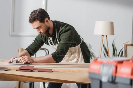 Repairman in apron sanding wooden plank near blurred toolbox in workshop 
