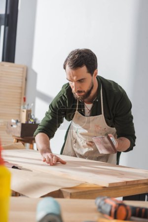 Bearded repairman holding sandpaper and checking wooden board in workshop 