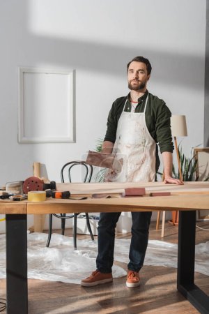 Artesano en delantal sosteniendo papel de lija y mirando a la cámara cerca de herramientas y tablero de madera en el taller 