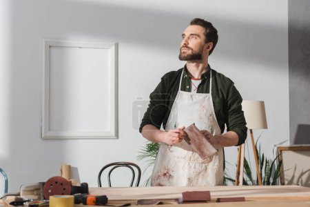 Photo for Bearded craftsman in apron holding sandpaper near blurred wooden board on table - Royalty Free Image