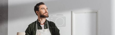 Photo for Dreamy craftsman in apron looking away in workshop, banner - Royalty Free Image