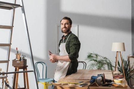 Restaurateur souriant en tablier tenant du papier de verre près des outils et du barde en bois en atelier 