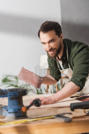 Positive restorer holding sandpaper near wooden board and sanding machine in workshop 
