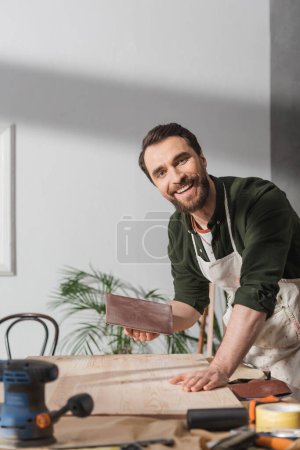 Lächelnder Tischler in Schürze hält Schmirgelpapier neben Brett und blickt in Werkstatt in die Kamera 