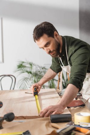 Restaurador en delantal midiendo tablero de madera con regla cerca de herramientas en taller 